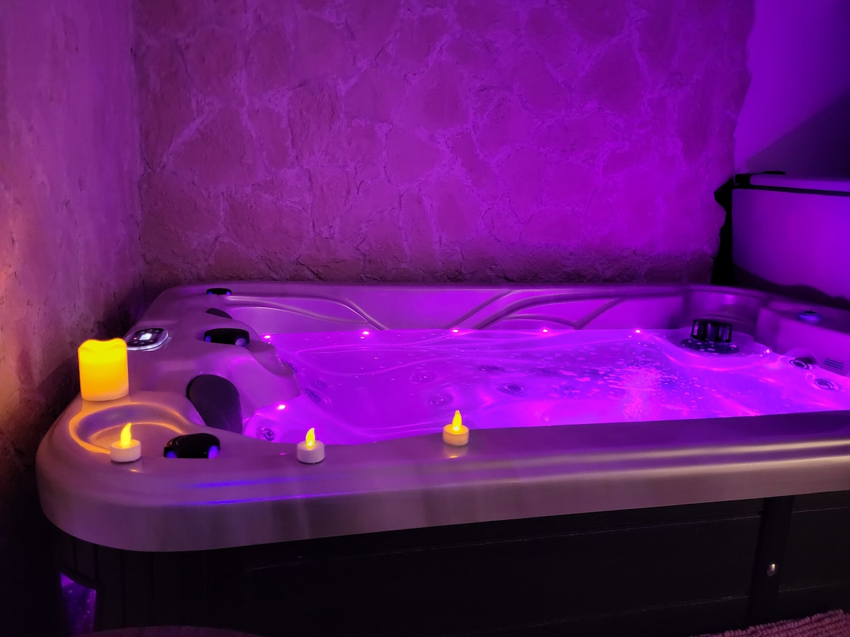 A soothing spa area is presented, featuring a vibrant, illuminated hot tub surrounded by soft purple lighting. Flickering candles add a calming element, while the textured stone wall provides an intimate setting. The spa area invites relaxation and tranquility.