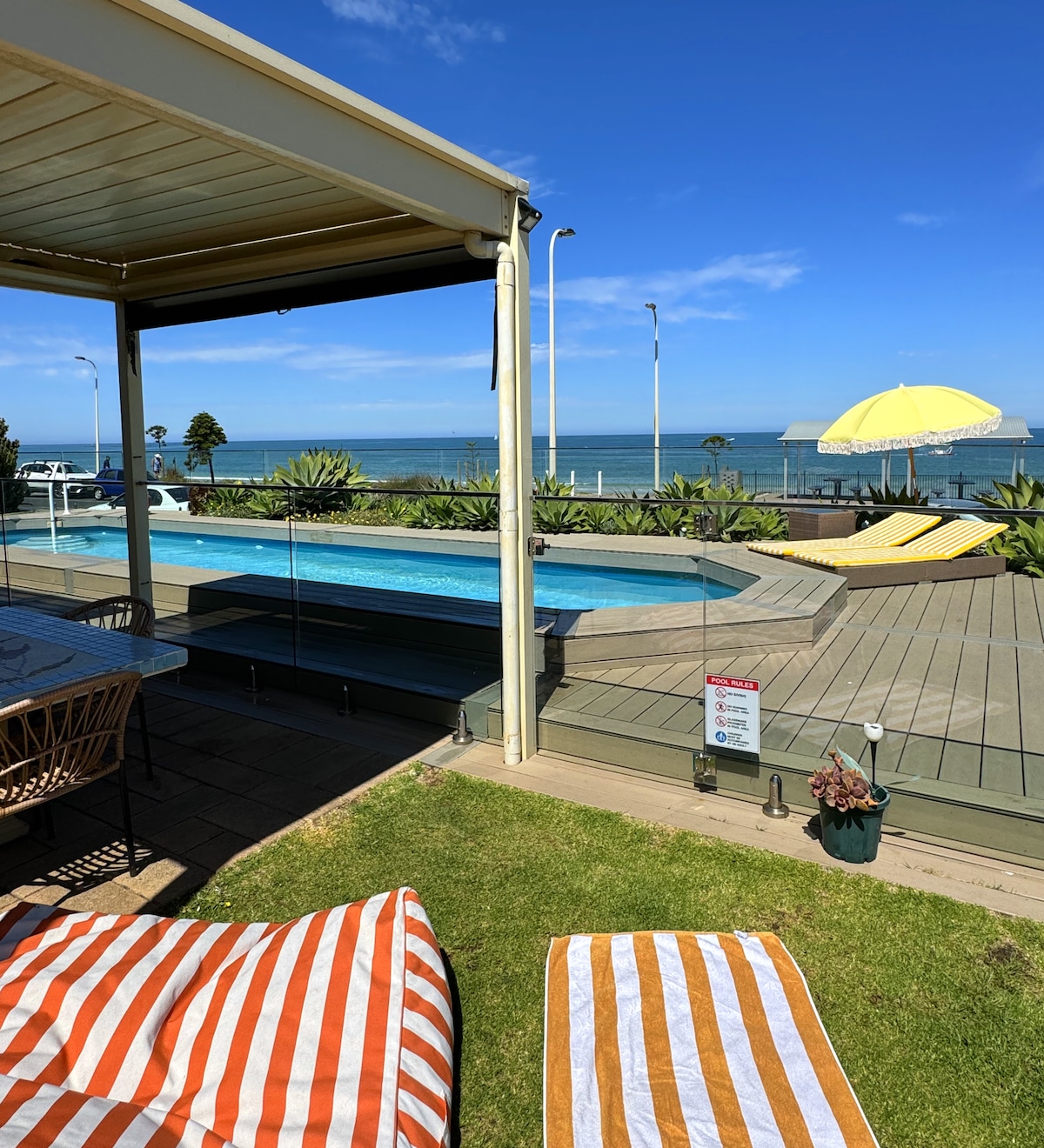 A private beachfront pool is surrounded by a spacious deck that offers unobstructed ocean views. Colorful loungers are positioned nearby, and an umbrella provides shade. The contemporary patio area includes a dining table, with lush greenery enhancing the coastal setting.