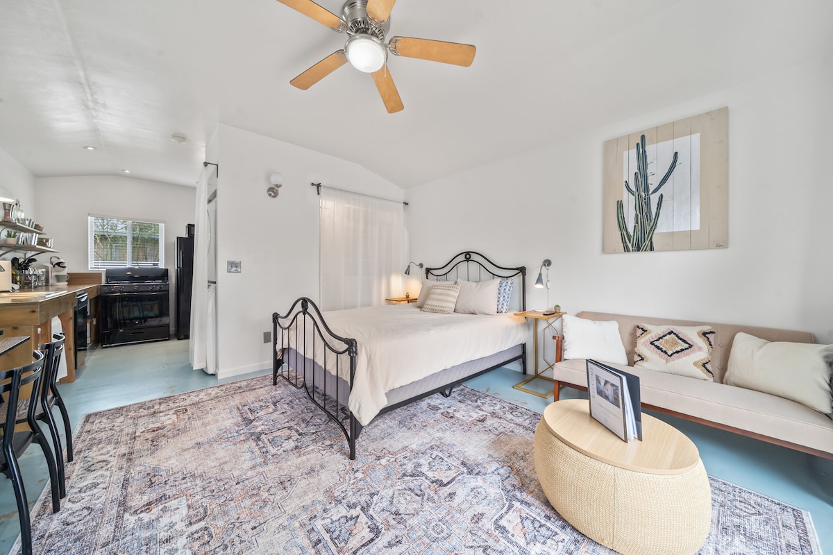 A spacious studio is showcased with a bed featuring a simple headboard and warm light fixtures on either side. A comfortable seating area is present, along with a large area rug beneath. Natural light is filtering through sheer drapes, contributing to a bright ambiance.
