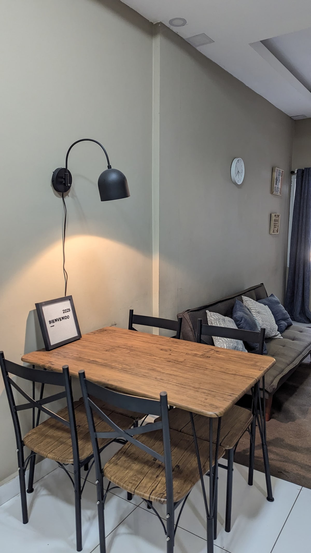 A dining area is depicted with a natural wooden table and three metal chairs. A stylish wall lamp casts soft light on the table. A cozy sofa with decorative pillows is positioned nearby, enhancing the space's functionality and comfort.