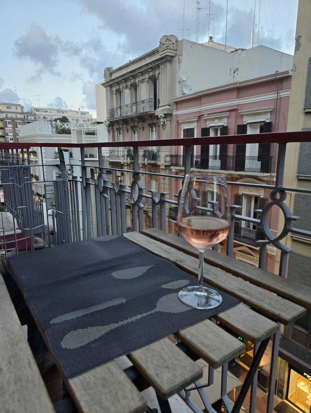 A small wooden table is situated on a balcony, accompanied by a glass of rose wine. The view reveals a charming street scene, featuring historic buildings with decorative facades. A metal railing provides an open feel while allowing for fresh air.