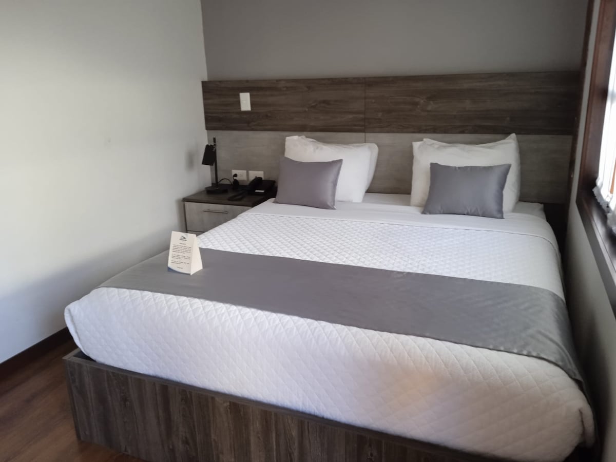 A cozy room features a spacious bed with gray accents and white bedding. Soft light enters through a nearby window, illuminating the inviting wood decor. A bedside table holds a lamp, and a phone is situated for easy access. A small sign is placed on the bed.