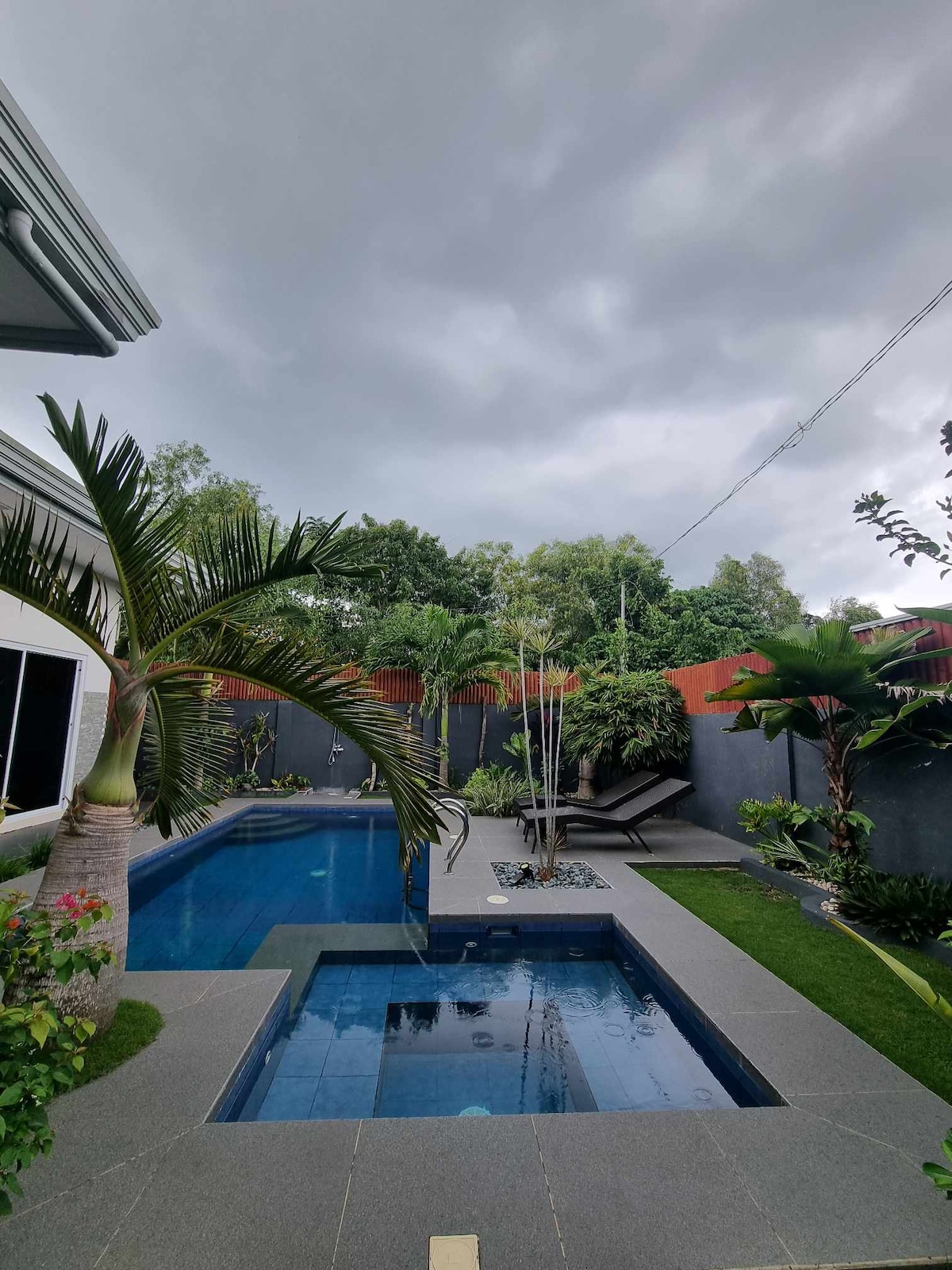 A private swimming pool features crystal-clear water, surrounded by a well-maintained tropical garden. Palms and other greenery enhance the vibrant atmosphere. The area is complemented by outdoor lounge chairs and a spa-like nook. The sky is overcast, adding a serene backdrop to the space.