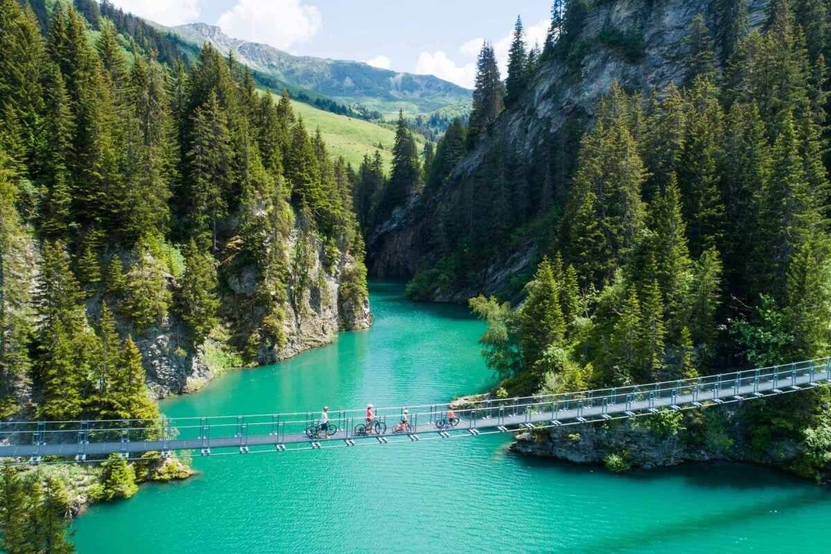 A suspension bridge spans across a vibrant turquoise river, surrounded by lush green trees and rocky cliffs. The mountains in the background are partially covered with greenery, creating a serene natural landscape. Small figures can be seen walking along the bridge.
