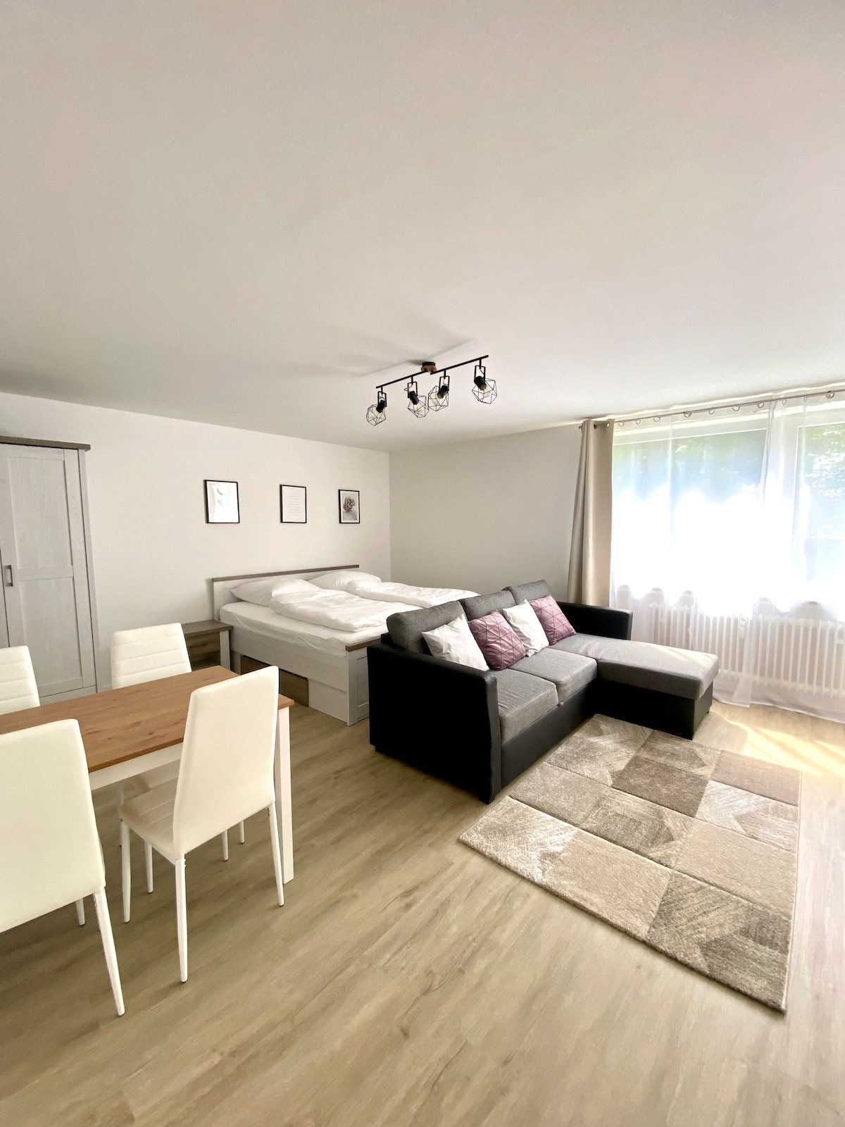 A cozy living space features a sleeping area with a comfortable bed and a stylish sectional sofa. A wooden dining table with four chairs is positioned nearby, while large windows allow natural light to fill the room. Neutral tones create a calm environment.