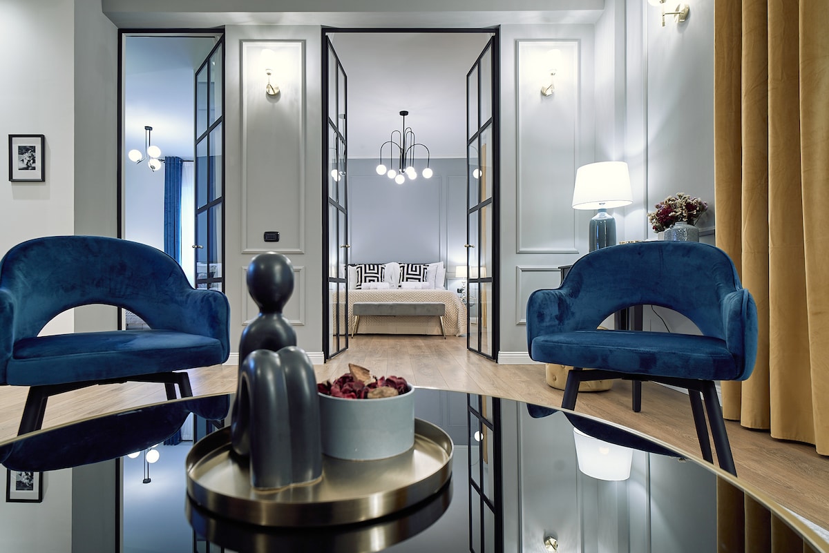 Two plush blue chairs are positioned around a round glass table, overlooking an elegant bedroom through double glass doors. Decor includes a modern lamp and a decorative tray containing a small bowl of dried fruits, enhancing the contemporary design.