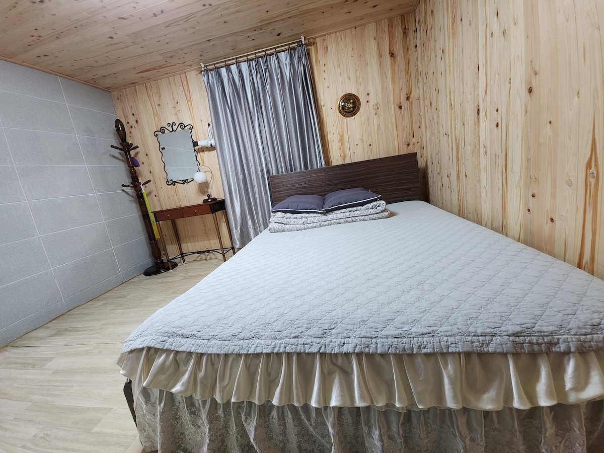 A spacious sleeping area features a large bed dressed in a light quilt, complemented by dark pillows. Wooden walls and a light-colored floor create a warm atmosphere. Natural light filters through elegant curtains, while a small mirror and desk are positioned nearby.