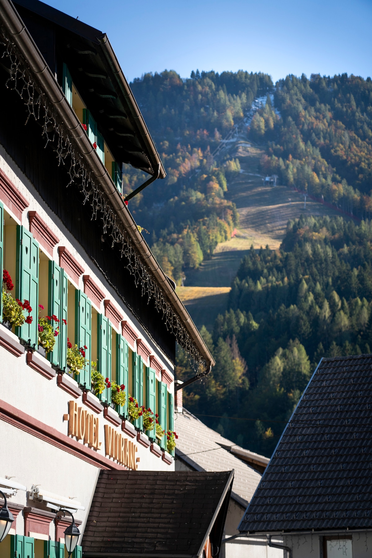 The exterior of the Vitranc Boutique Hotel showcases its charming façade, adorned with green window shutters and flower boxes. A backdrop of lush hills and ski slopes complements the inviting structure, highlighting the connection to nature and outdoor activities.