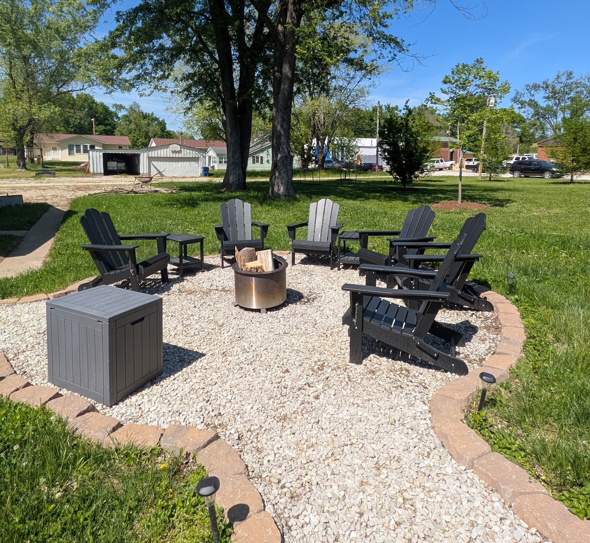 An outdoor firepit area is set with six black Adirondack chairs arranged in a circle around a stone firepit. A gray storage box sits nearby, and a gravel surface provides a foundation, surrounded by lush green grass and shaded by trees.