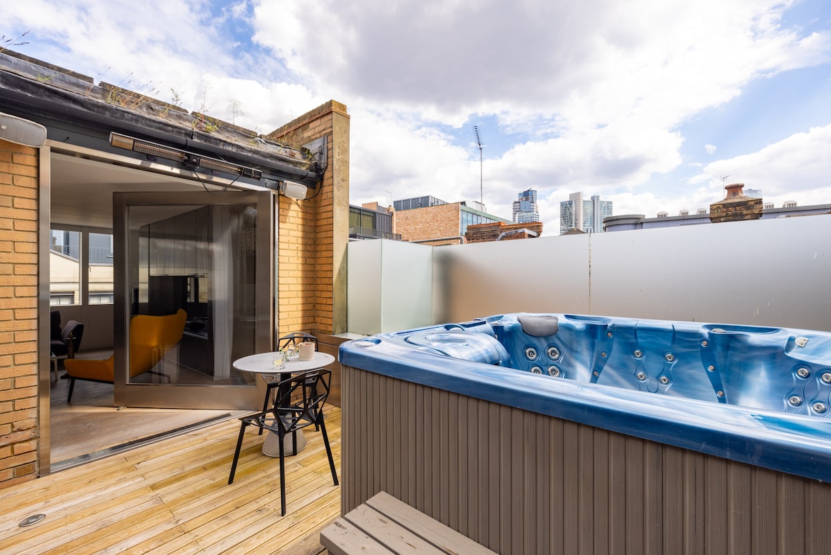 An outdoor terrace features a bubbling hot tub surrounded by wooden decking. A small table with two chairs is positioned nearby, offering a cozy spot to relax. The urban skyline is visible in the background under a partly cloudy sky.