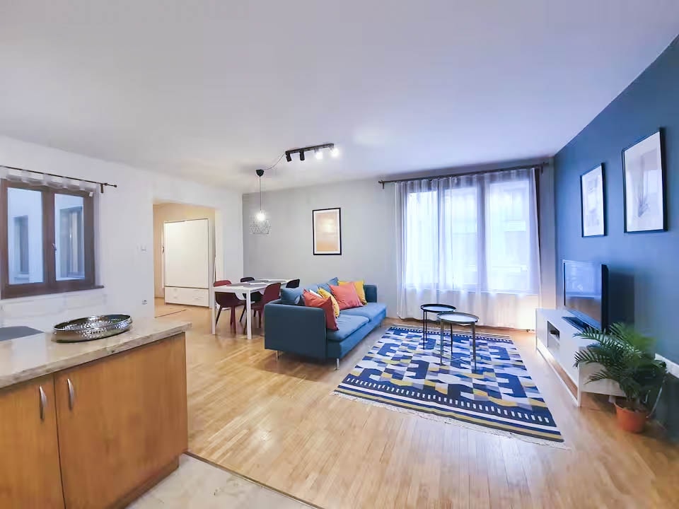 The living area is furnished with a comfortable sofa adorned with colorful pillows, a coffee table, and a flat-screen TV. Natural light fills the space through large windows, illuminating the contemporary decor on the walls. A dining table is visible in the background, enhancing the inviting atmosphere.