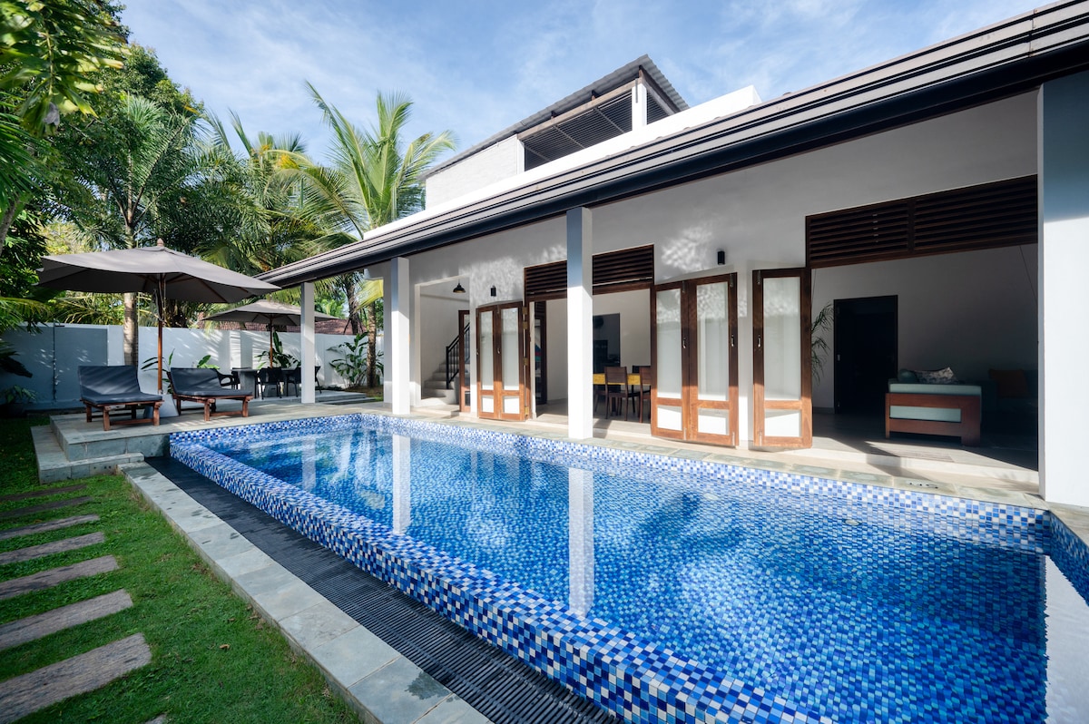 The pool area features a rectangular pool adorned with mosaic tiles, surrounded by a sun deck equipped with loungers and umbrellas. Large bi-fold doors provide a seamless connection to the indoor living space and dining area, framed by lush greenery.