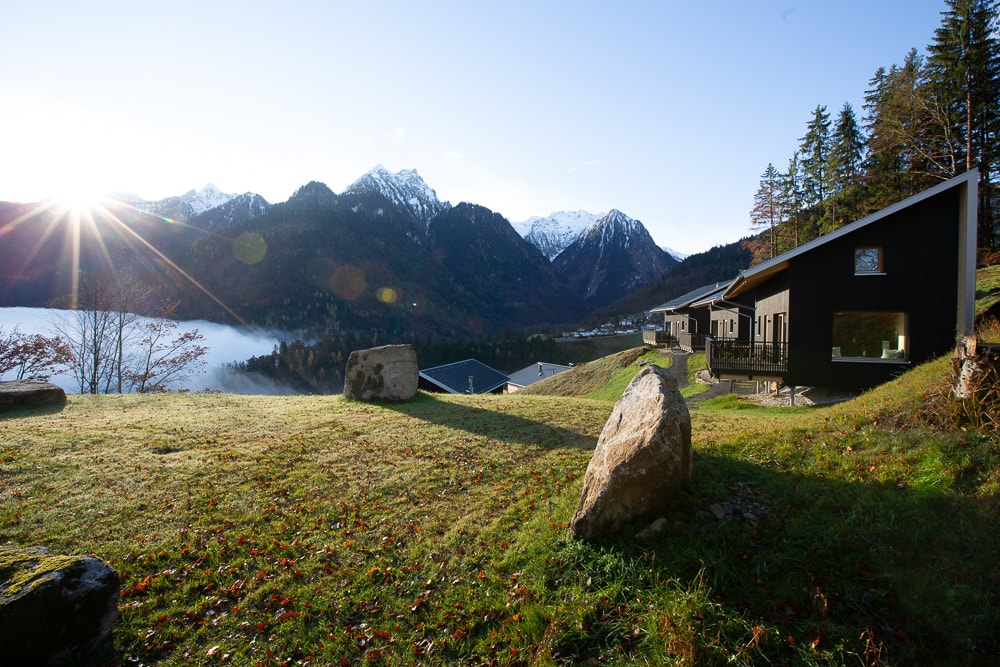 A scenic outdoor view captures the modern cabins nestled among rolling hills, surrounded by mountains and greenery. Sunlight streams down from the left, illuminating the landscape. Large boulders and grass create a natural setting, enhancing the peaceful ambiance of the location.