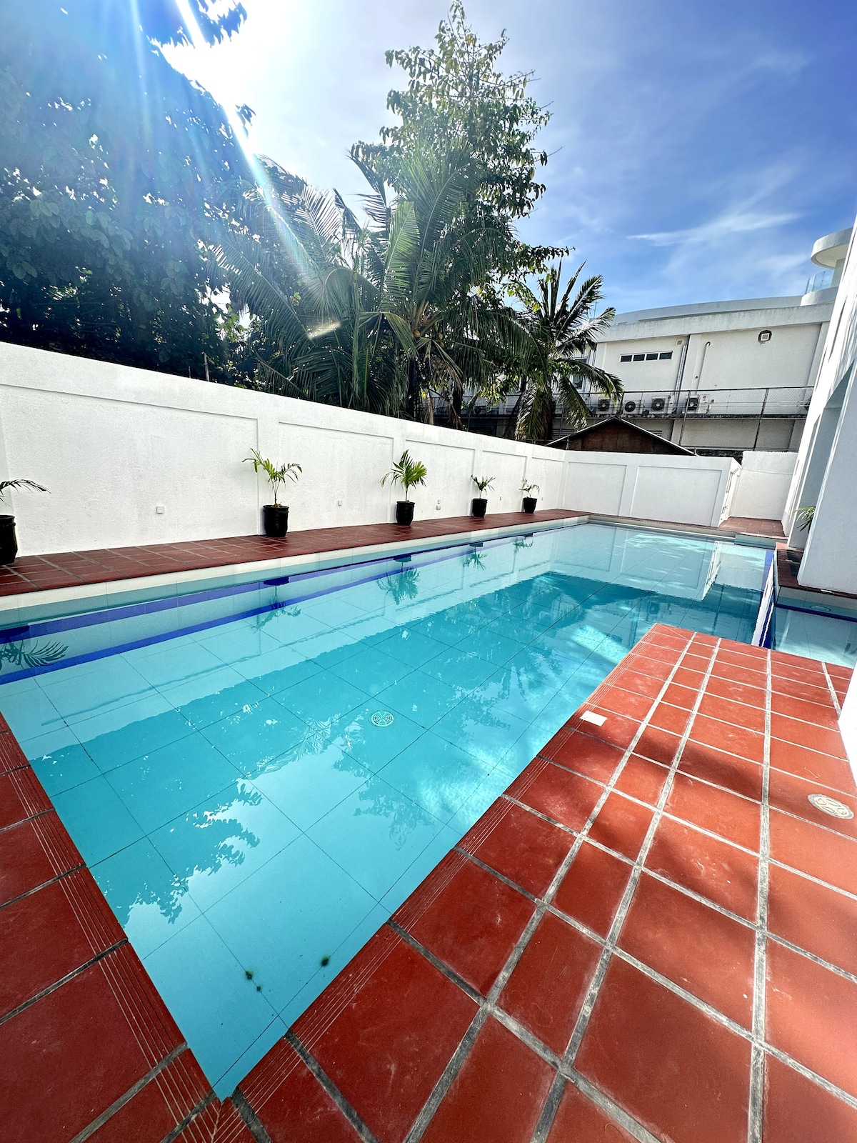 A clear blue pool is framed by a red-tiled deck and bordered by potted plants. Lush tropical foliage is visible in the background, while the surrounding walls provide a sense of privacy, creating a serene environment for relaxation.