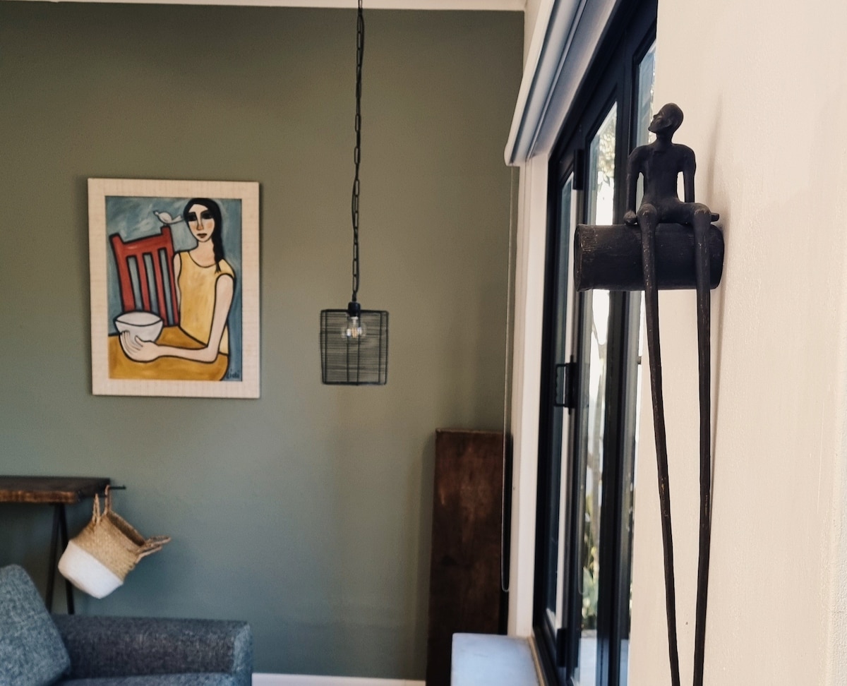 A lounge area features a modern grey sofa beside a large window, allowing natural light to fill the space. A framed artwork depicting a seated figure with a bowl is displayed on the green wall, while an industrial-style pendant light hangs from the ceiling.