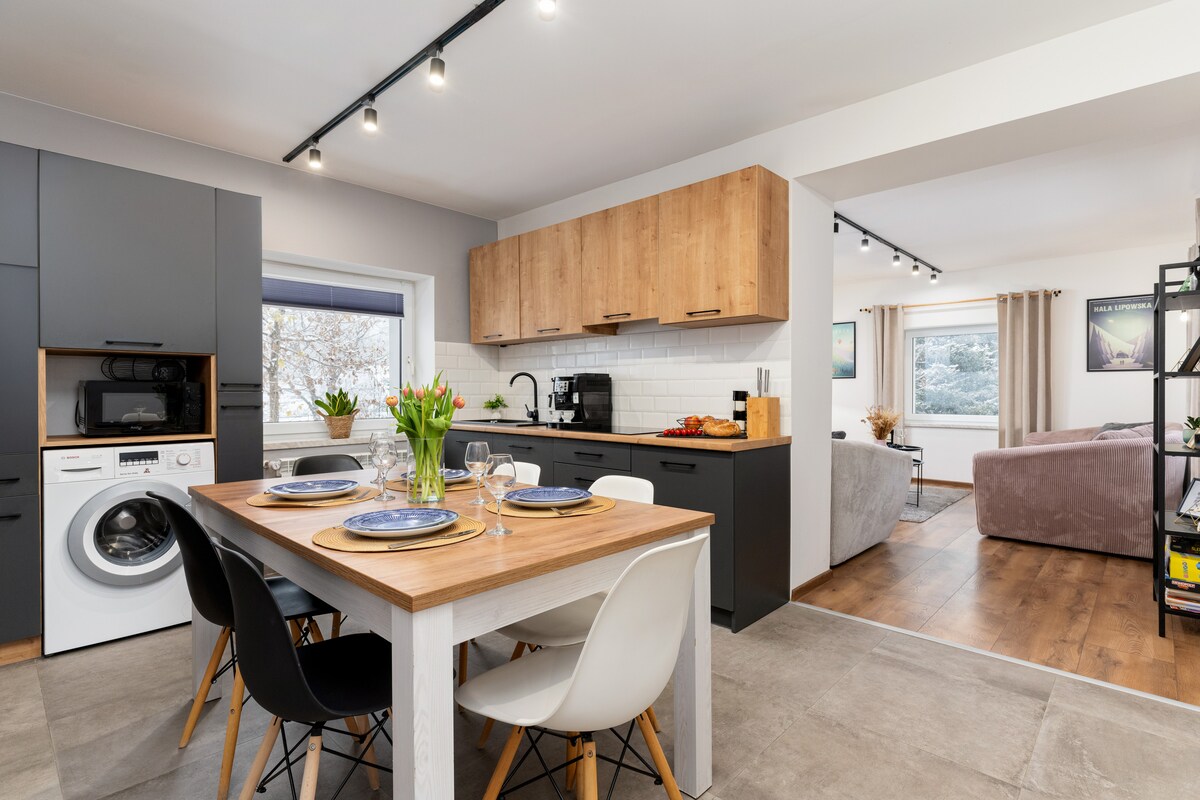 A modern kitchen space is highlighted by a wooden dining table set for four, accented with elegant dishes. Nearby, a well-equipped kitchen features sleek cabinetry and essential appliances, including a washing machine. Natural light filters through the window, illuminating the inviting living area in the background.