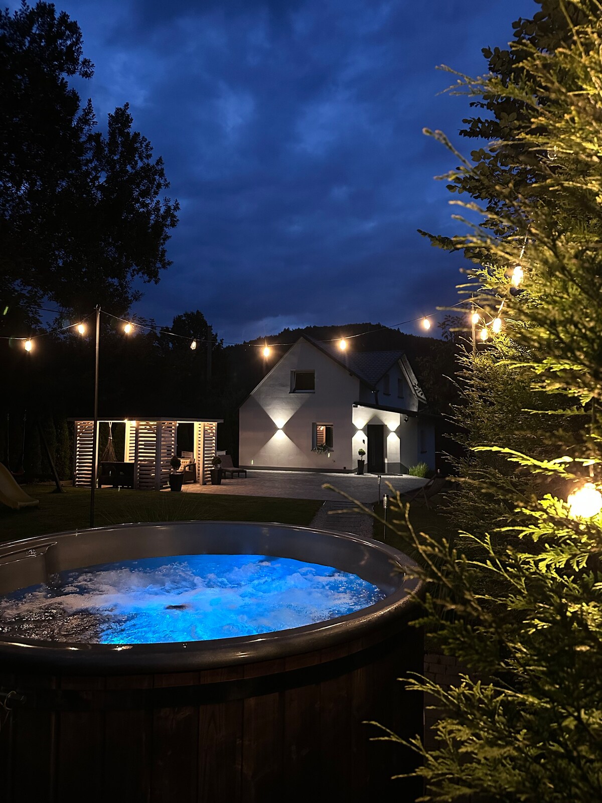 A warm-lit exterior showcases a wooden hot tub with blue illumination, set against a backdrop of cloudy skies. The house is visible in the distance, framed by trees and decorative string lights, creating a cozy ambiance for evening relaxation.