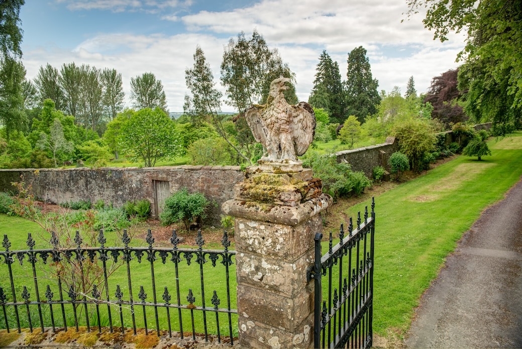 An ornate stone eagle statue is positioned atop a pillar, overlooking a lush, landscaped garden. A low stone wall surrounds the garden, which features a variety of trees and greenery, creating a serene and spacious atmosphere. A pathway runs alongside, inviting exploration.