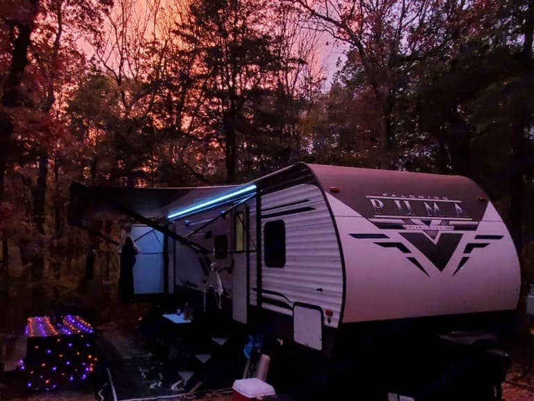 The travel trailer is positioned amid trees, featuring a retractable awning and exterior lighting. The scene is illuminated by a vibrant sunset sky, enhancing the tranquil outdoor setting. An outdoor mat is visible beneath the camper, contributing to the inviting atmosphere.