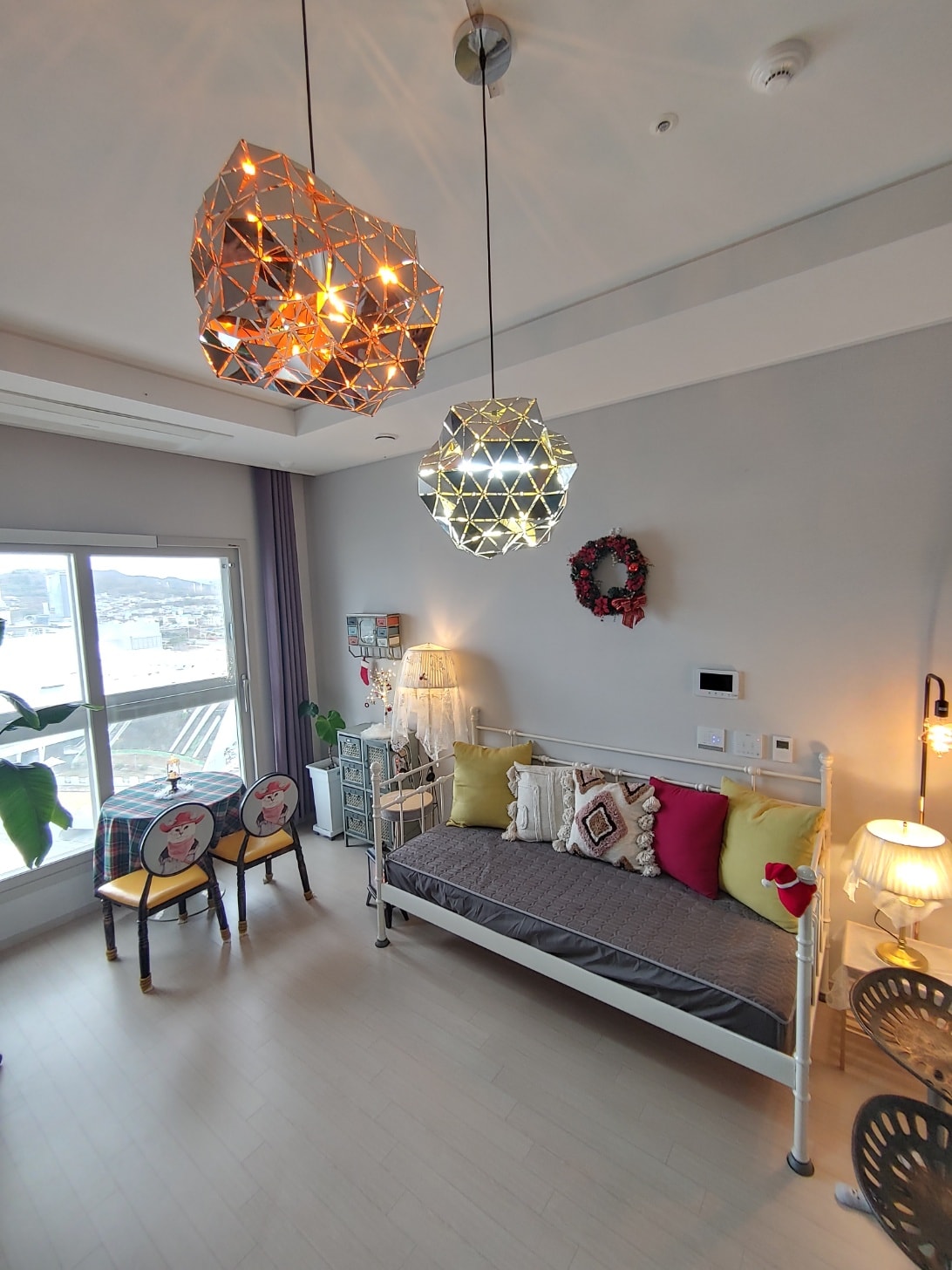 A bright living area features modern light fixtures casting warm light. A spacious daybed is adorned with colorful cushions, and a small dining table with two chairs is visible. Large windows provide ample natural light and a view of the surroundings.