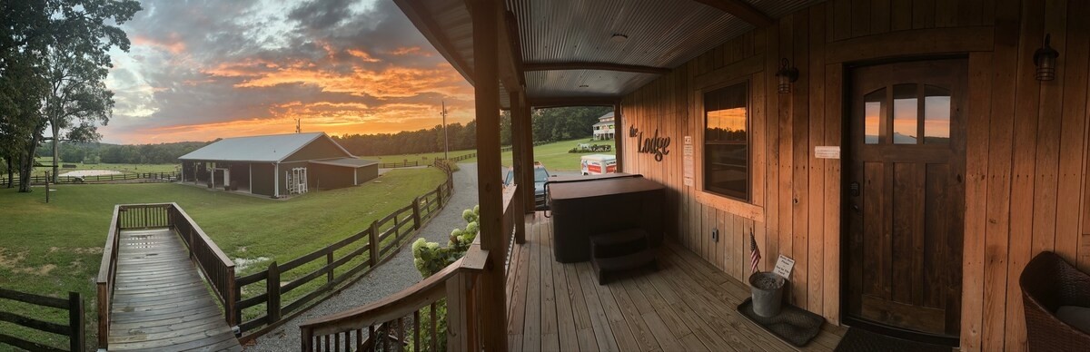 The front porch features a private hot tub and wooden railing, with scenic views of the countryside. A sunset casts vibrant colors across the sky, while the spacious lawn and fenced area create a serene setting. The rustic cabin entrance is visible to the right.