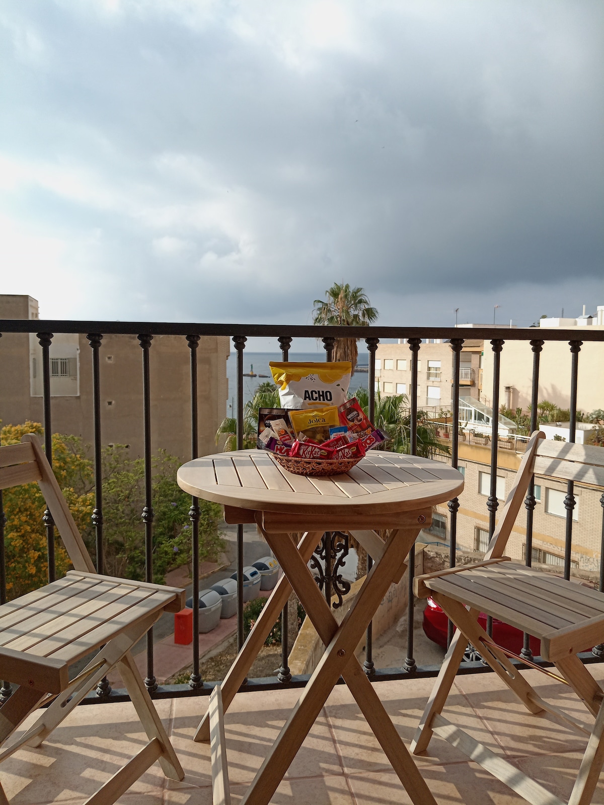 A small, round wooden table is positioned on the balcony, accompanied by two folding chairs. A basket filled with snacks is visible on the table. The backdrop features a view of nearby buildings and greenery, with hints of the sea in the distance.