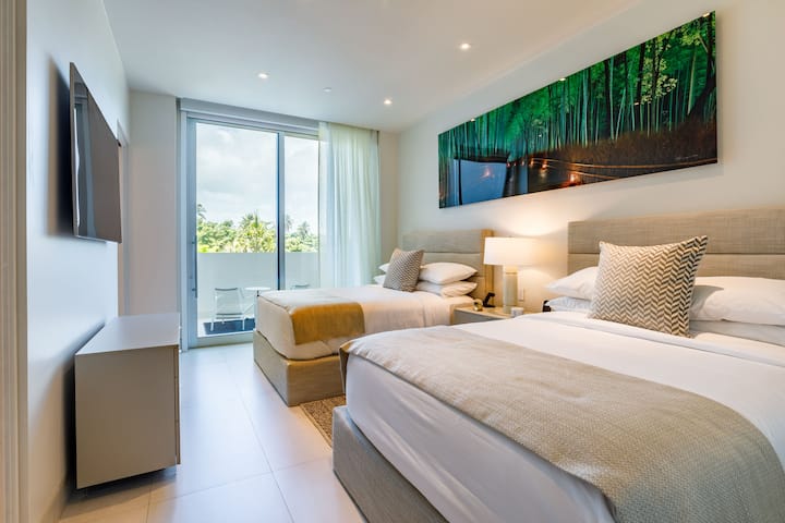 This serene bedroom features two queen-size beds dressed in luxurious linens, complemented by a calming nature-inspired artwork. Sliding doors lead to a private balcony, inviting natural light and fresh air.