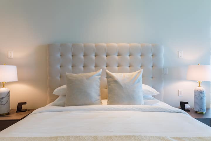 The bed showcases a plush tufted headboard, crisp white bedding, and cozy pillows. Flanked by chic marble-based lamps, the minimalist yet elegant decor ensures comfort and style.