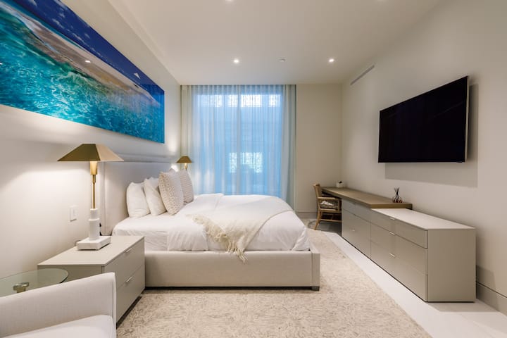 The king-size bed with neutral tones and luxury linens is complemented by a stunning ocean-themed artwork. A cozy armchair and a sleek dresser complete the room.