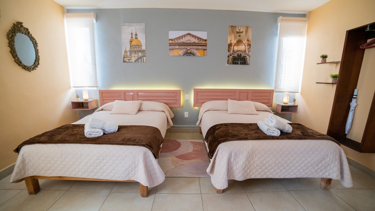 The sleeping area features two spacious beds, each arranged with soft white linens and brown throws. The room is illuminated by natural light from windows dressed in light sheer curtains. Wall art depicting architectural details enhances the atmosphere, while a round mirror adds depth to the space.
