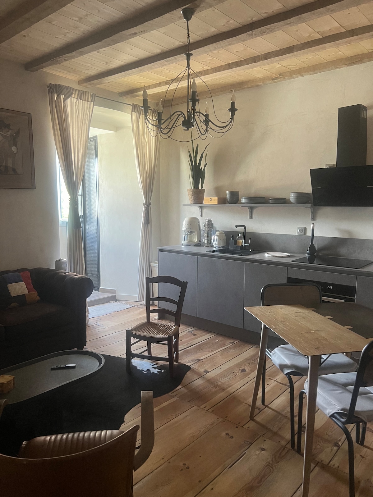 A cozy kitchen and living area features wooden beams overhead and a large window that allows natural light to fill the room. A dining table is set near a simple black couch, and a chandelier hangs above, providing a touch of elegance.