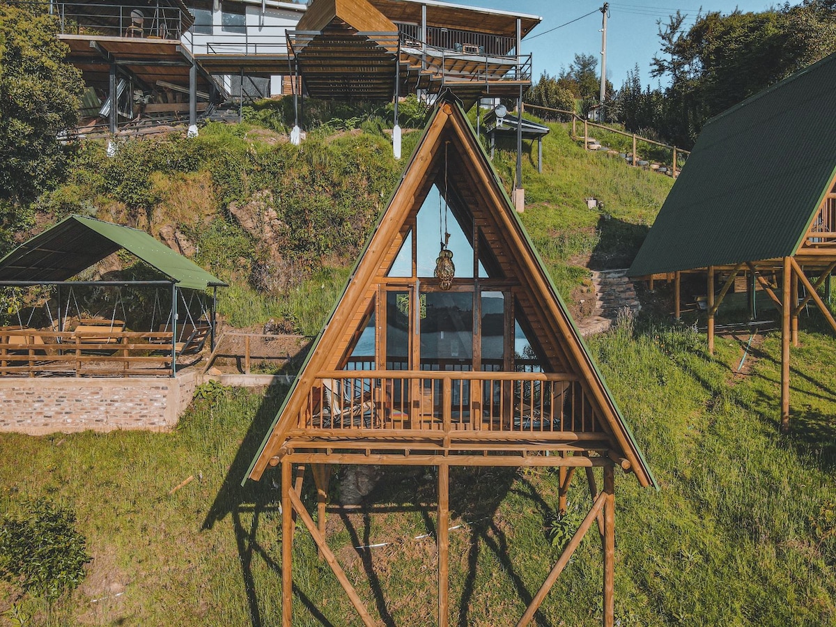An A-frame cabin stands prominently on a hillside, showcasing large glass windows that invite natural light. Nearby wooded areas and open green spaces enhance the setting. A separate outdoor structure provides a communal space, complementing the cabin's rustic design.