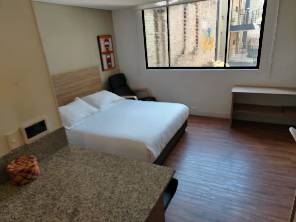 A sleeping area is presented with a spacious bed featuring white linens. Natural light enters through a large window, lighting the wooden floor. A small desk and a chair are positioned in the room, while a wall displays a subtle art piece.