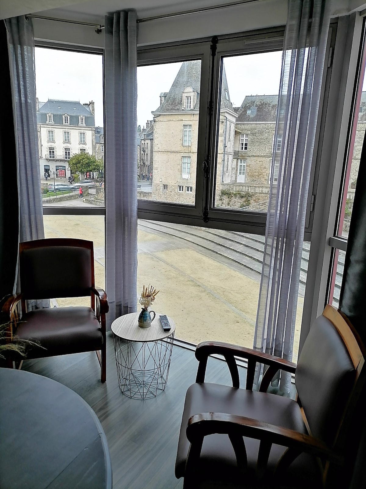 A corner seating area is presented with two wooden chairs positioned beside a stylish metallic side table. Large windows with sheer curtains frame a view of the historic architecture outside, allowing natural light to illuminate the space.