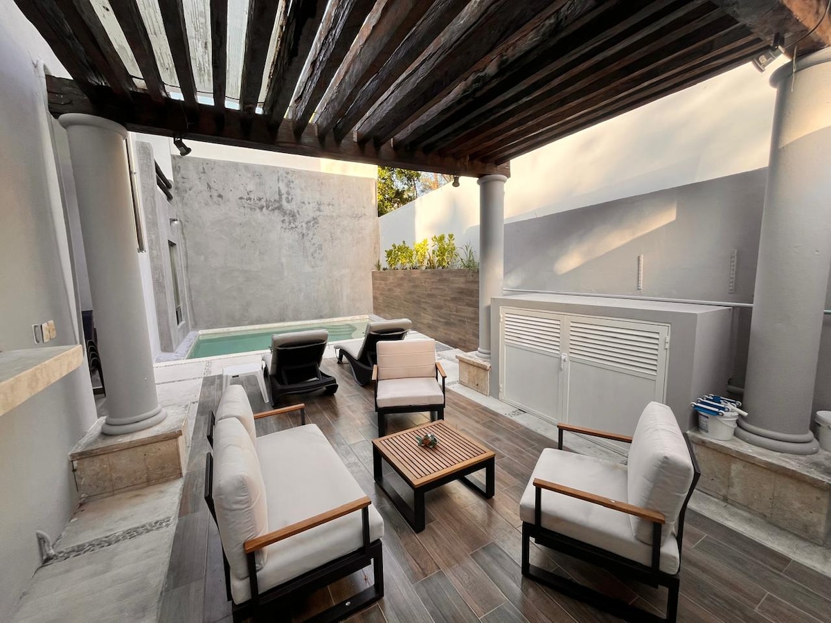 An outdoor patio area features a wooden slatted roof providing shade over a set of four cushioned armchairs and a coffee table. A small pool is visible in the background, with stone walls outlining the space.