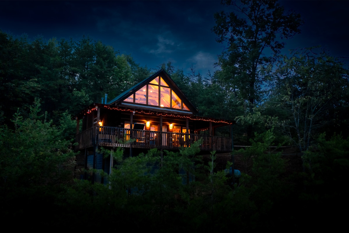 The cabin is framed by trees, illuminated softly from within. Its large windows highlight a triangular roof structure, while string lights enhance the inviting exterior ambiance. The surrounding greenery creates a sense of privacy and seclusion, complementing the natural setting.