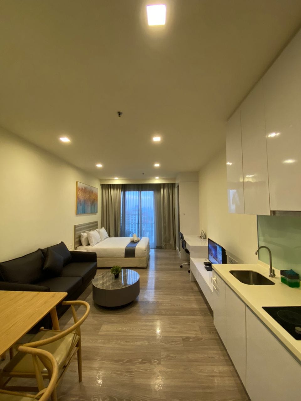 A cozy living space features a sofa and dining table positioned near a well-equipped kitchen. A well-lit area is dominated by a spacious bed, with soft linens and decorative pillows. The large window offers city views, while neutral tones create a calm environment.