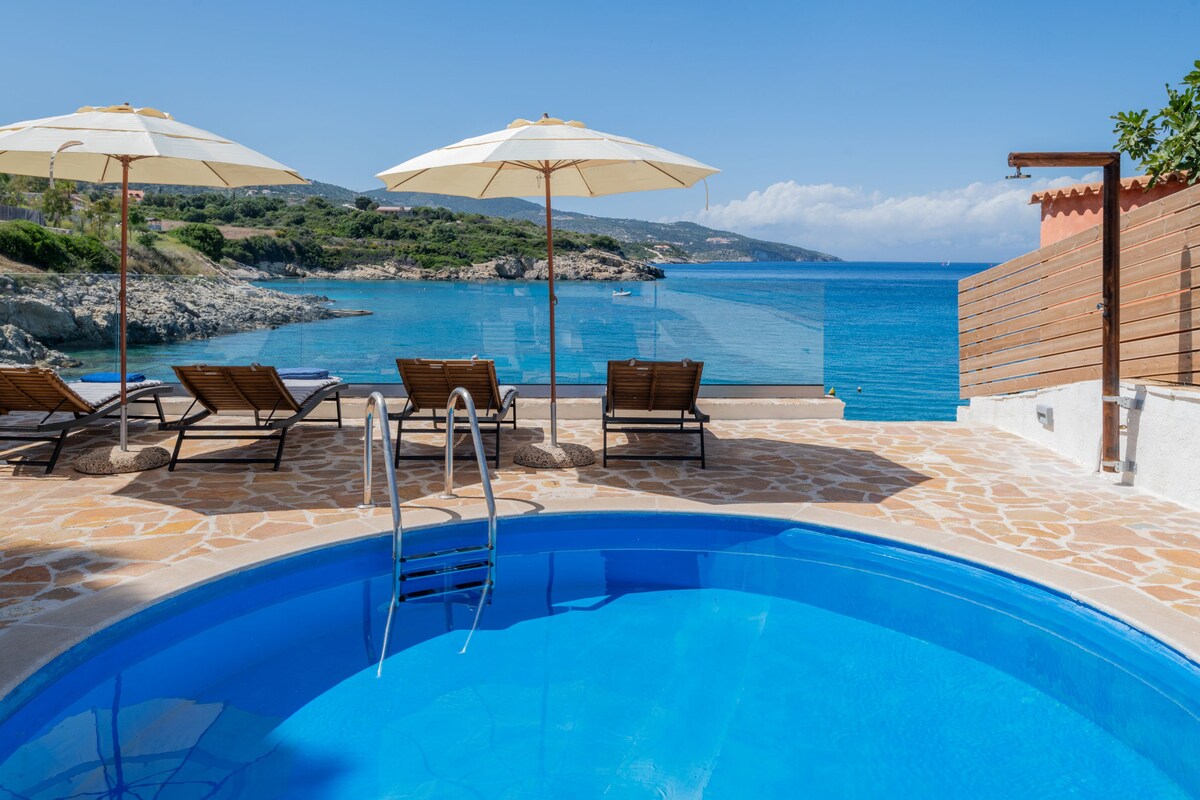 A serene outdoor space features a private pool surrounded by sun loungers under large umbrellas. The view overlooks calm, clear waters and a rocky coastline, with green hills in the distance under a bright blue sky.