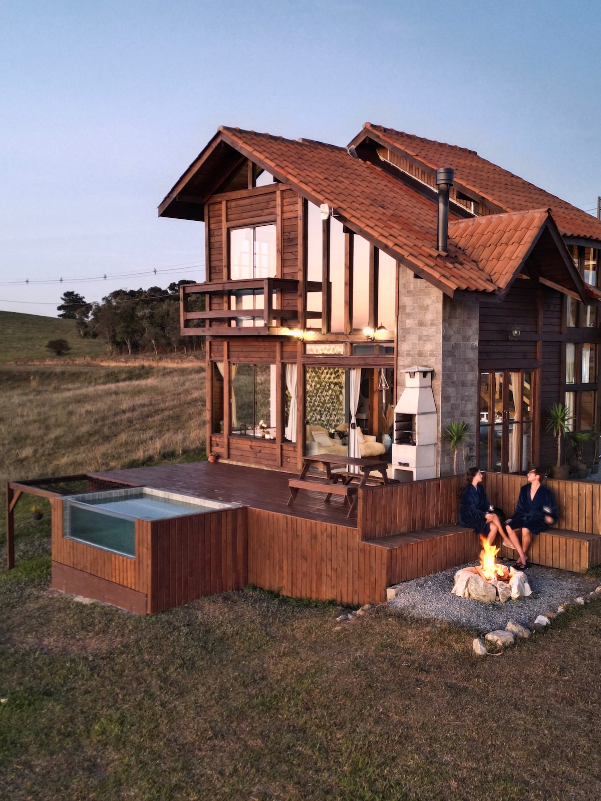 A wooden cabin features a warm exterior with a tiled roof and large windows, revealing a cozy interior. A private deck surrounds a heated jacuzzi and a fire pit, all set against expansive grassy fields under a soft evening sky.