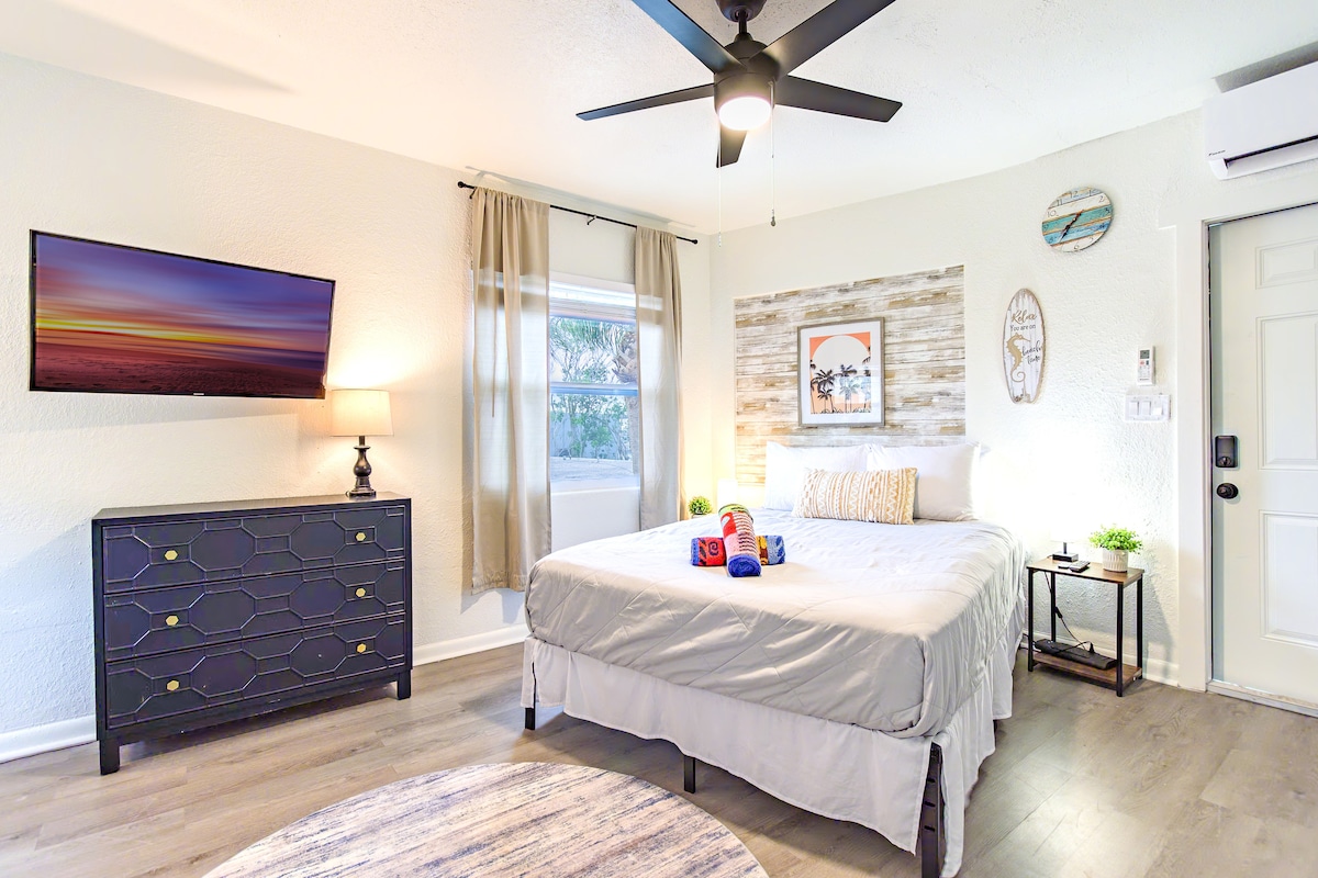 A bright room features a spacious bed with neutral linens. A dark wood dresser with a decorative lamp and a television are positioned nearby. A small table with potted plants complements the decor, while natural light enters through curtains next to the window.