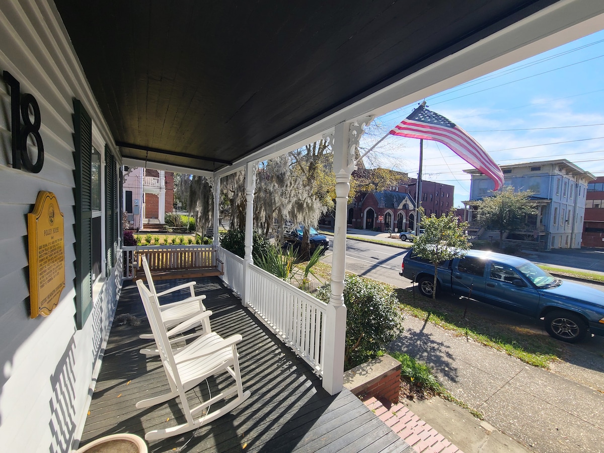 A charming porch is featured with white rocking chairs positioned for relaxation. A flag is displayed prominently on a nearby street, while the surrounding greenery and historic buildings are visible in the background, enhancing the inviting outdoor space.