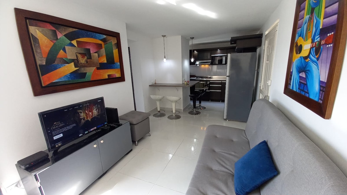 The living area features a gray sofa facing a television stand, with a vibrant piece of artwork on the wall. A small dining bar with two stools is visible, leading into a modern kitchen equipped with appliances. Bright lighting enhances the spacious atmosphere.