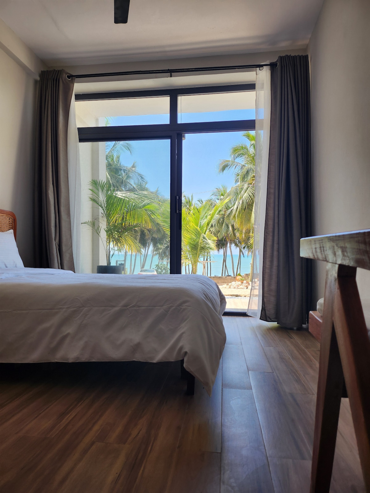 A serene bedroom features a spacious bed with crisp white linens. Large glass doors open to a view of palm trees and the ocean, allowing natural light to fill the room. Wooden flooring adds warmth to the tranquil atmosphere.