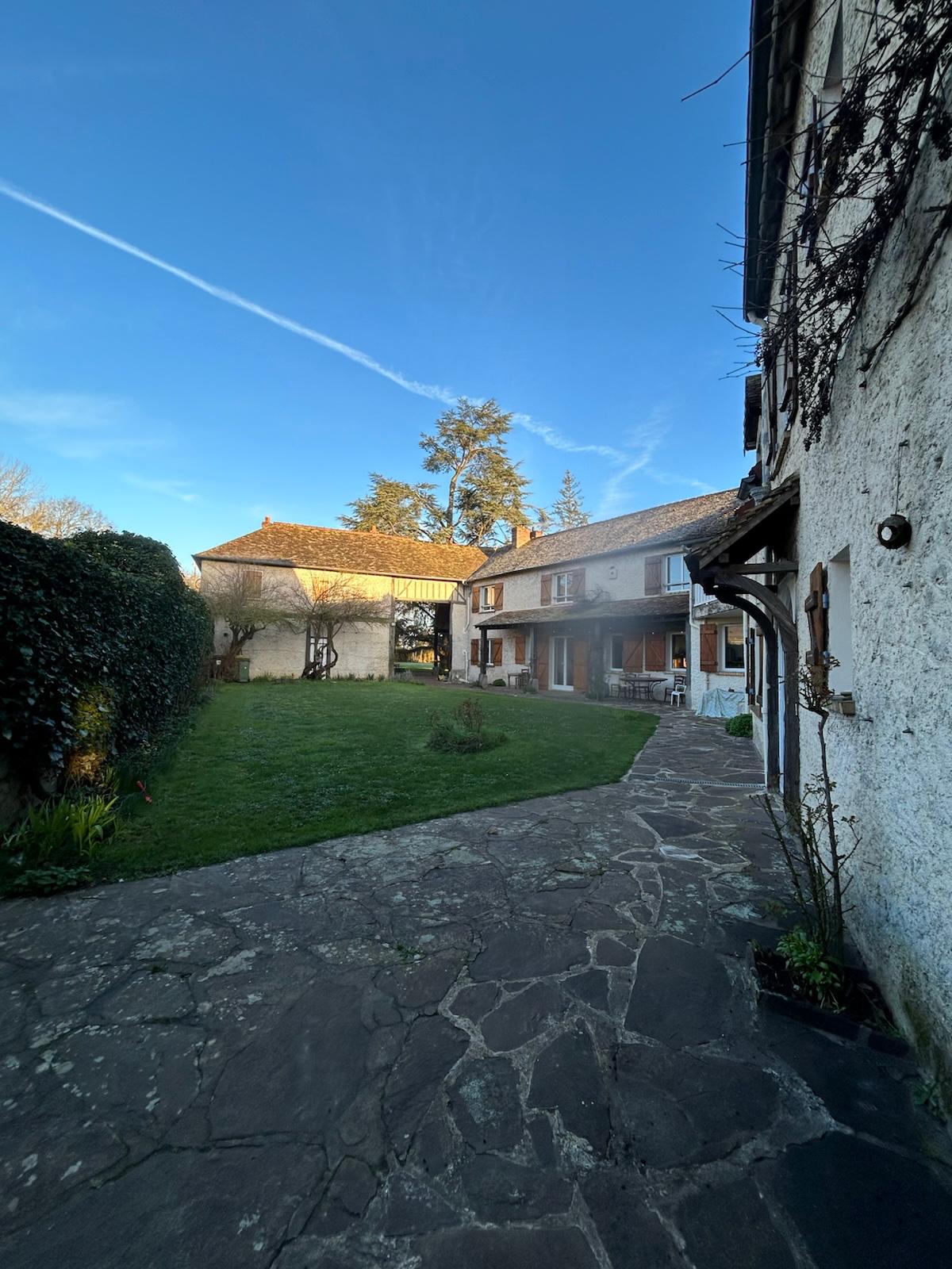 A serene outdoor area featuring a stone pathway leading between two charming buildings. Lush greenery lines the pathway and a clear blue sky is visible above, enhancing the peaceful atmosphere. The architecture reflects a blend of rustic charm and modern comfort.
