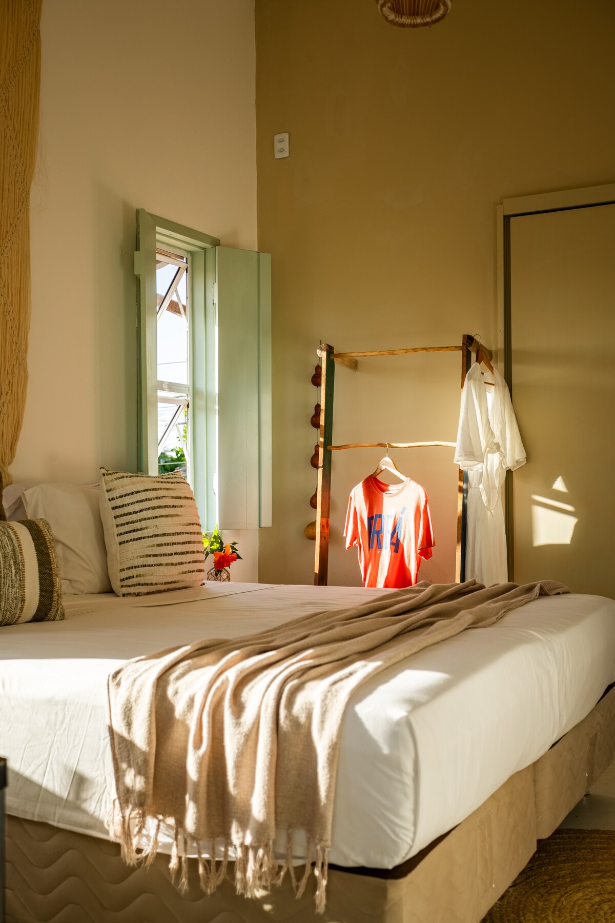 A bright and airy bedroom is presented, featuring a spacious bed with a light-colored duvet. A clothing rack displays a casual outfit, while natural light streams through a window adorned with green shutters, creating a warm and inviting ambiance.