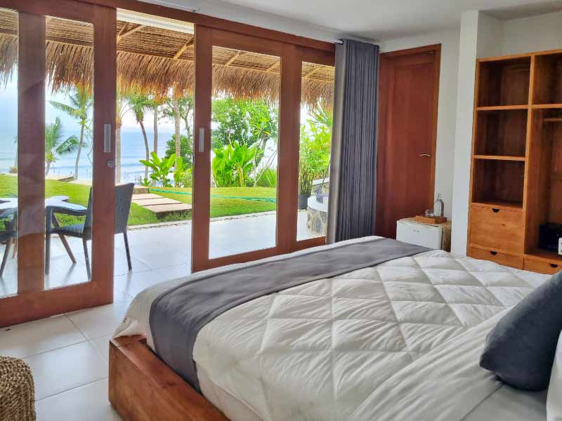 A spacious sleeping area features a comfortable bed with a quilted coverlet. Large glass doors provide a view of the lush garden and ocean beyond. Natural light enhances the room's warm tones, with wooden furniture and a neatly arranged bookshelf complementing the décor.