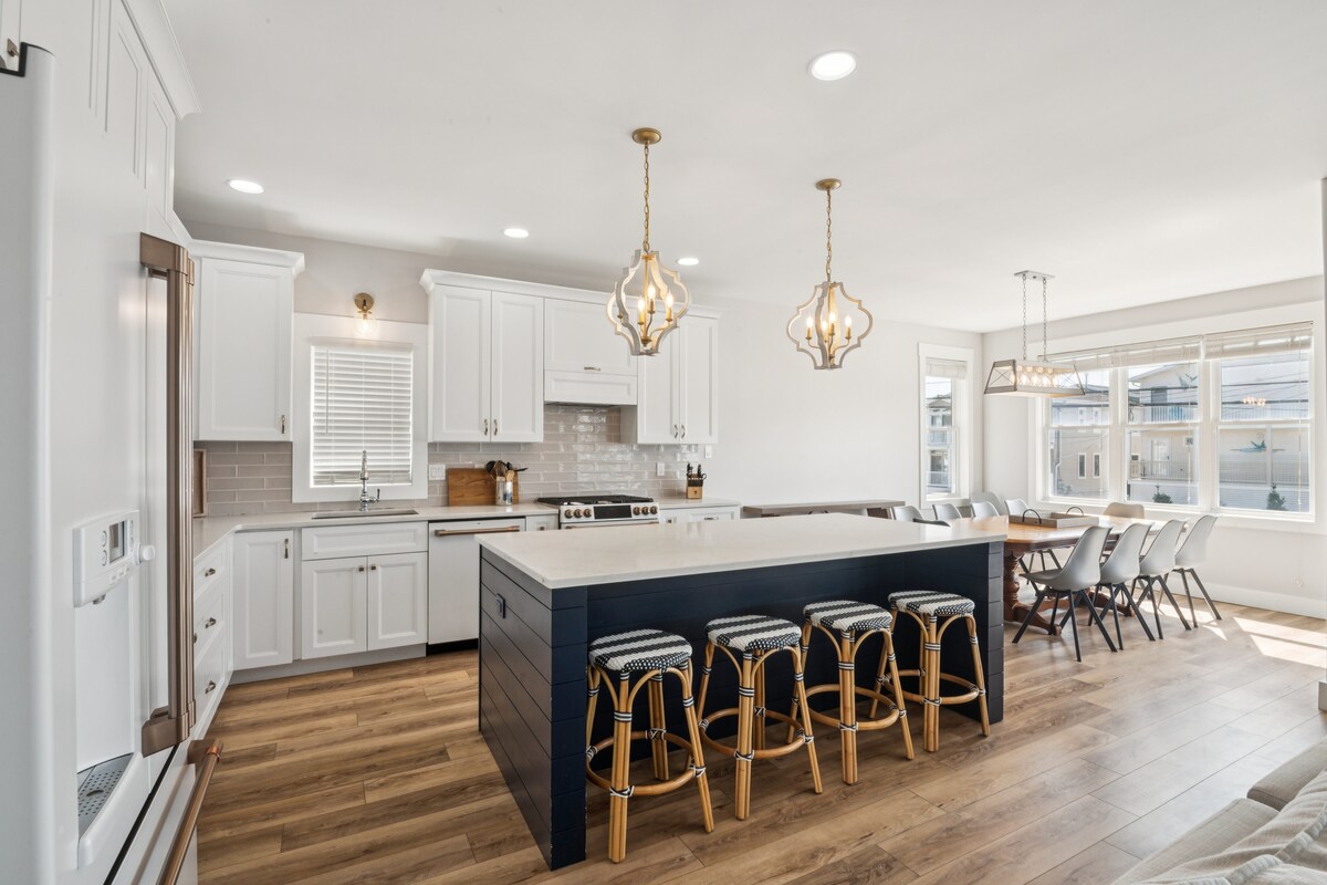 A bright kitchen and dining area is presented with modern finishes. An island with seating for six is complemented by stylish pendant lighting. White cabinetry and countertops offer a clean aesthetic, while abundant natural light fills the space through nearby windows.