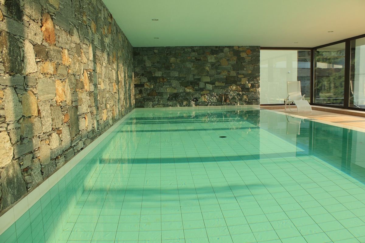 An indoor swimming pool features light blue waters reflecting the room's ceiling. A stone wall adds texture to the space, while a single lounger rests beside the pool, inviting relaxation. Large windows allow natural light to illuminate the area.