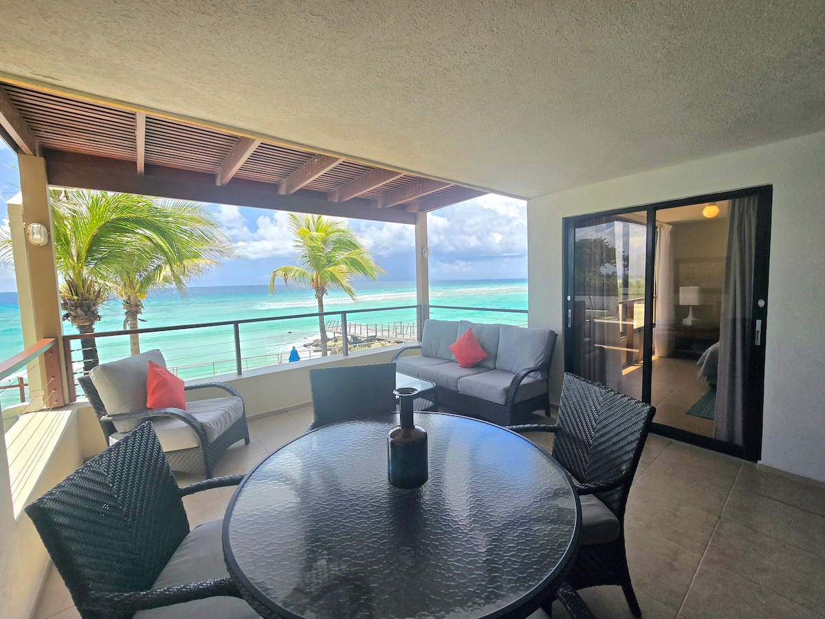 A partially covered balcony is equipped with a round dining table and four woven chairs. A comfortable seating area includes a sofa and two pillows, with views of the beach and the Caribbean Sea visible through large glass doors and palm trees swaying in the breeze.