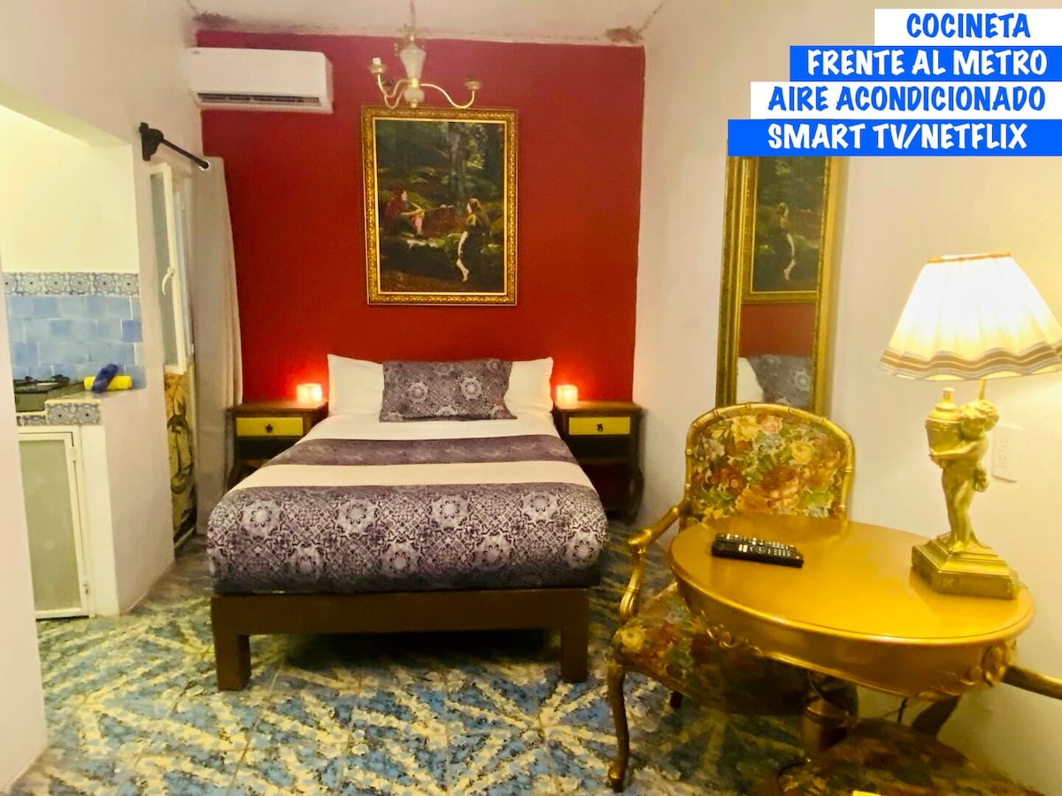 A cozy room features a spacious bed with decorative bedding, complemented by a vintage-style chair and gold-framed artwork on the wall. Soft lighting is provided by bedside lamps, and air conditioning is visible in the ceiling, with a kitchenette seen in the corner.